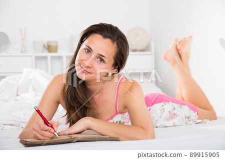 Portrait of young smiling woman lounging in bed and writing to diary Portrait of young smiling woman lounging in bed and writing to diary 89915905