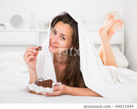 Adult woman secretly eating chocolate under sheets in bed 89915944