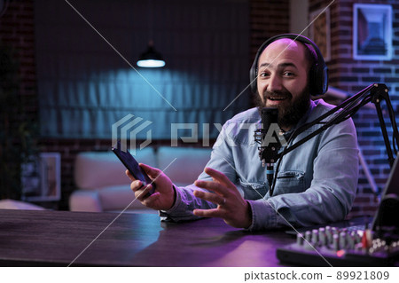 POV of male influencer reviewing smartphone product on camera, filming podcast video in studio with neon lights. Online vlogger doing mobile phone recommendation on internet livestream. POV of male influencer reviewing smartphone product on camera, filming podcast video in studio with neon lights. Online vlogger doing mobile phone recommendation on internet livestream. 89921809