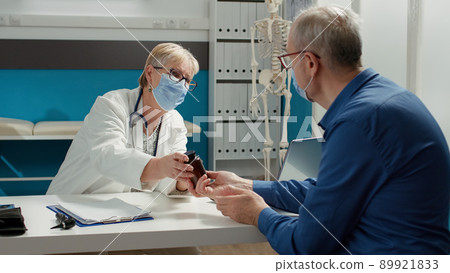 General practitioner and patient with face mask talking about pills bottle in medical office. Physician giving prescription treatment and medicine to retired man, to cure disease. General practitioner and patient with face mask talking about pills bottle in medical office. Physician giving prescription treatment and medicine to retired man, to cure disease. 89921833