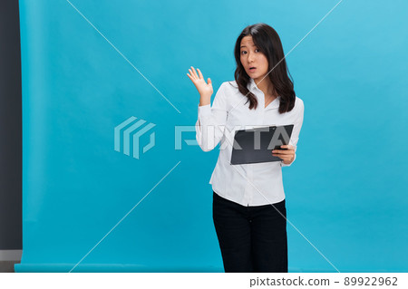 Shocked cute Asian businesswoman in classic office dress code holds folder tablet tells secret posing isolated on over blue studio background. Cool business offer. Job interview concept Shocked cute Asian businesswoman in classic office dress code holds folder tablet tells secret posing isolated on over blue studio background. Cool business offer. Job interview concept 89922962