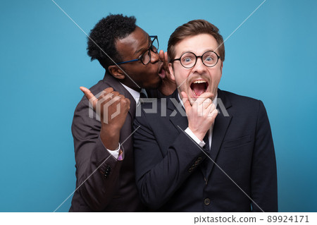 Two shocked young men coworkers whisper gossip and tell secret behind his hand sharing news. 89924171
