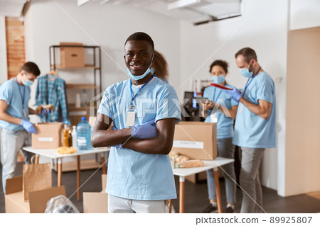 Portrait of smiling african american young male volunteer in blue uniform, protective mask and gloves standing with arms crossed. Team sorting, packing food stuff in cardboard boxes Portrait of smiling african american young male volunteer in blue uniform, protective mask and gloves standing with arms crossed. Team sorting, packing food stuff in cardboard boxes 89925807