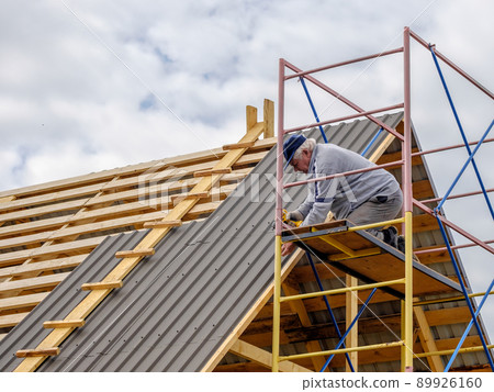 Senior builder man with a screwdriver screwing a roofing sheet to the roof. 89926160