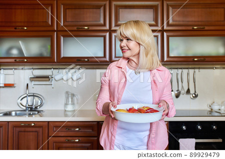 Beautiful mature woman with prepared plate of apples for baking in home kitchen Beautiful mature woman with prepared plate of apples for baking in home kitchen 89929479