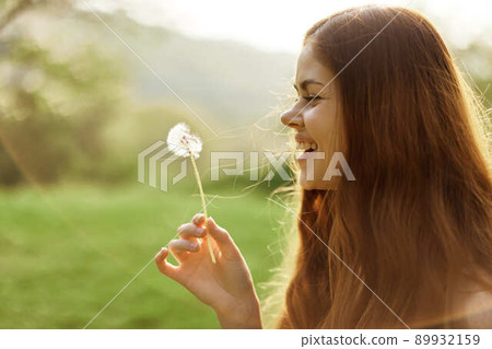 Portrait of a young woman in profile with a dandelion flower in her hand blowing on it and smiling against the green summer grass in the setting sun in nature 89932159