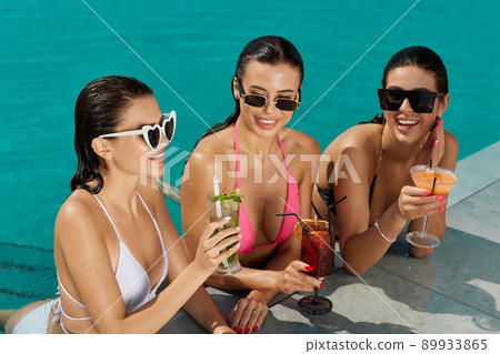 Three glamorous ladies in sunglasses sunbathing, having fun on swimming pool deck in summer. Front view of smiling women in bikinis relaxing, drinking cocktails in blue pool water. Concept of relax. 89933865