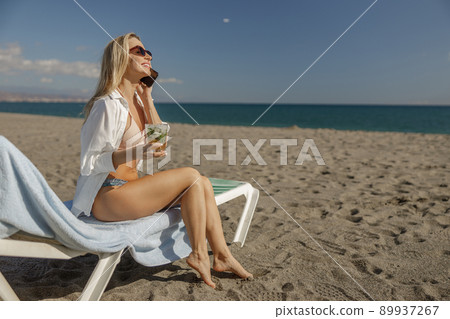 Full length shot of lady in bikini and shirt on deck chair having talk on a phone 89937267