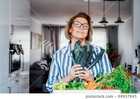 Smiling woman holding fresh kale from her healthy food delivery. Wooden box with vegetables. Support Local farmer food. Start of a healthy life, weight loss concept. Online home food delivery. 89937845