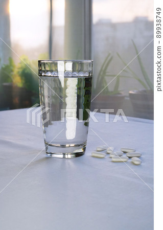 A transparent glass of water and white medicinal tablets next to it on a table in the living room. Side view. Eye-level angle A transparent glass of water and white medicinal tablets next to it on a table in the living room. Side view. Eye-level angle 89938749