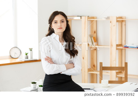 Smiling mature professional businesswoman with arms crossed sitting on the desk in office. 89939392