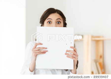 Young woman with surprised eyes peeking out from behind paper poster. Businesswoman holding big white banner in modern office. Young woman with surprised eyes peeking out from behind paper poster. Businesswoman holding big white banner in modern office. 89939407