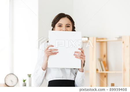 Young woman with surprised eyes peeking out from behind paper poster. Businesswoman holding big white banner in modern office. Young woman with surprised eyes peeking out from behind paper poster. Businesswoman holding big white banner in modern office. 89939408