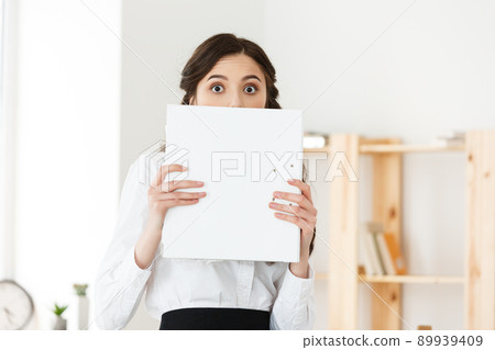 Young woman with surprised eyes peeking out from behind paper poster. Businesswoman holding big white banner in modern office. Young woman with surprised eyes peeking out from behind paper poster. Businesswoman holding big white banner in modern office. 89939409
