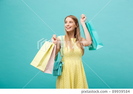 Lifestyle Concept: Portrait of shocked young attractive woman in yellow summer dressposing with shopping bags and looking at camera over blue background. Lifestyle Concept: Portrait of shocked young attractive woman in yellow summer dressposing with shopping bags and looking at camera over blue background. 89939584