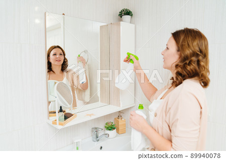 an attractive housewife washes the bathroom mirror with a special glass cleaner. 89940078