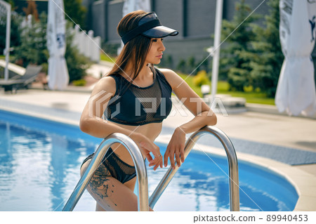 Portrait of a woman having rest and posing near a swimming pool. Dressed in a black swimsuit and sun visor. 89940453