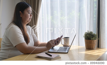 Side view chubby asian woman sitting in front of laptop computer and using mobile phone at home 89948890