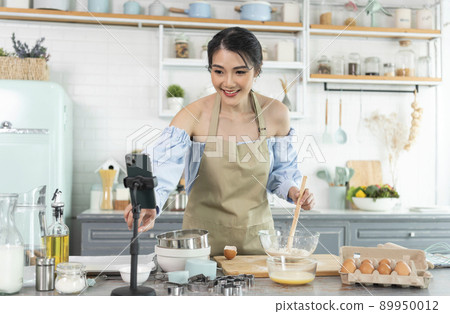 Asian woman food blogger cooking cake in front of smartphone camera while recording vlog video and live streaming at home in kitchen. 89950012