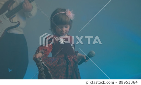 Little girl on stage in vintage dress, she sings into microphone and dances, her father plays an electric guitar. Color music is shining and smoke is billowing. Family time, creativity and hobbies 89953364