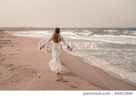 Model in boho style in a white long dress and silver jewelry on the beach. Her hair is braided, and there are many bracelets on her arms. Model in boho style in a white long dress and silver jewelry on the beach. Her hair is braided, and there are many bracelets on her arms. 89953890