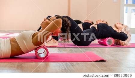A group athletic women doing pilates or yoga on pink mats in front of a window in a beige loft studio interior. Teamwork, good mood and healthy lifestyle concept. 89953999