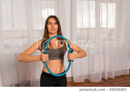 young woman with magic pilates ring, fitness instructor in Sportswear Leggings and Tops, stretching in the gym, on a yoga mat near the large window on a sunny day, female fitness yoga routine concept 89956801
