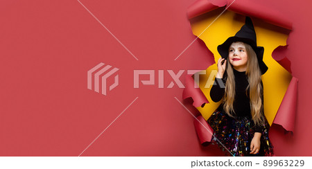 Laughing funny child girl in a witch costume of halloween looking, smiling and scares through a hole of red, yellow paper background. Copyspace 89963229