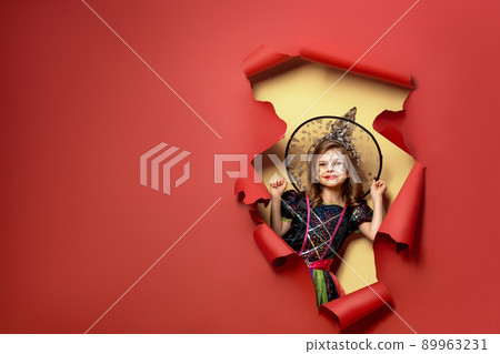 Laughing funny child girl in a witch costume of halloween looking, smiling and scares through a hole of red, yellow paper background. Copyspace 89963231