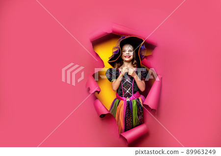 Laughing funny child girl in a witch costume of halloween looking, smiling and scares through a hole of red, yellow paper background. Copyspace Laughing funny child girl in a witch costume of halloween looking, smiling and scares through a hole of red, yellow paper background. Copyspace 89963249