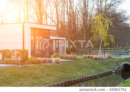 Modern country house with the large lawn and a wooden fence. In front of the house there is a covered terrace with a lounge zone. On the lawn there is a trees and flowerbeds. It is sunny. Outside. Modern country house with the large lawn and a wooden fence. In front of the house there is a covered terrace with a lounge zone. On the lawn there is a trees and flowerbeds. It is sunny. Outside. 89963762