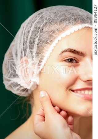 Plastic surgery and beauty concept. Professional cosmetician examining and touching the face of the beautiful young caucasian smiling woman in a clinic of esthetic cosmetology Plastic surgery and beauty concept. Professional cosmetician examining and touching the face of the beautiful young caucasian smiling woman in a clinic of esthetic cosmetology 89964127