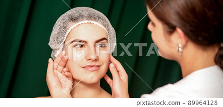 Plastic surgery and beauty concept. Professional cosmetician examining and touching the face of the beautiful young caucasian smiling woman in a clinic of esthetic cosmetology Plastic surgery and beauty concept. Professional cosmetician examining and touching the face of the beautiful young caucasian smiling woman in a clinic of esthetic cosmetology 89964128