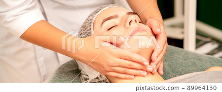 Facial treatment massage. Beautiful young caucasian woman with perfect skin receiving face and neck massage at a beauty spa Facial treatment massage. Beautiful young caucasian woman with perfect skin receiving face and neck massage at a beauty spa 89964130