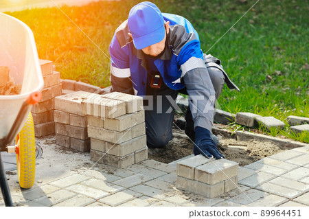 Bricklayer in work clothes sits on sidewalk and lays out paving slabs. Sight of working man in open 89964451