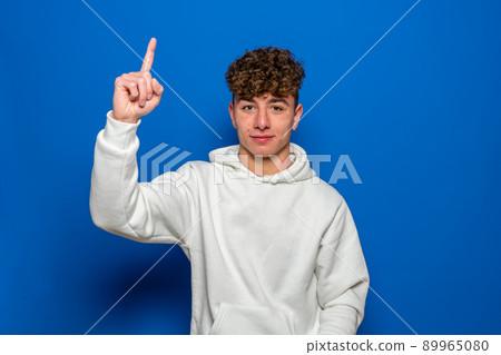 Caucasian young man with curly hair dressed in a white sweatshirt pointing the index finger up, isolated on blue studio background. Caucasian young man with curly hair dressed in a white sweatshirt pointing the index finger up, isolated on blue studio background. 89965080
