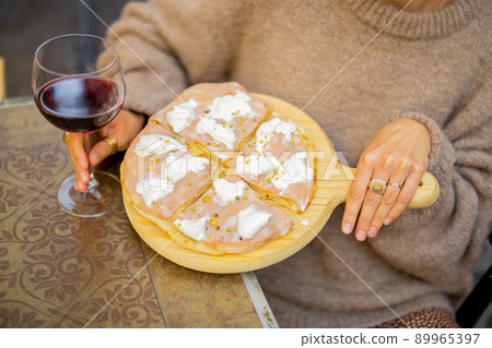 Woman holding wooden board with small pizza and glass of red wine at outdoor restaurant Woman holding wooden board with small pizza and glass of red wine at outdoor restaurant 89965397
