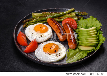 A delicious nourishing breakfast with fried eggs, sausages, asparagus, tomatoes, avocado, spices and herbs 89966185