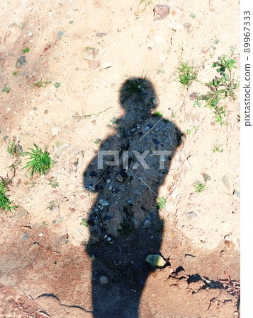 Long shadow of a person in jacket with hood on dry sand with growing green grass and lots of stones and rocks Long shadow of a person in jacket with hood on dry sand with growing green grass and lots of stones and rocks 89967333