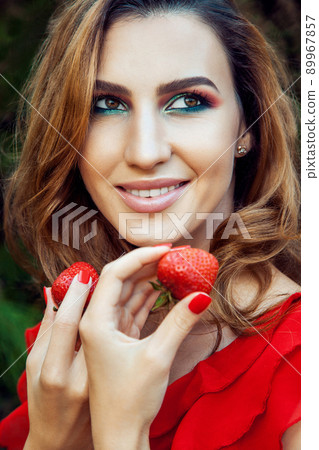 Young beautiful happy funny girl with red dress and makeup holding strawberry in summertime in the park. healthy lifestyle, diet beauty and happiness concept. 89967857