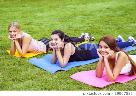 Group of beautiful satisfied healthy slimy young friends have rest after doing exersices on the green grass in park, lie on mat and head up with hands. Outdoor, sport lifestyle, yoga time, wellness Group of beautiful satisfied healthy slimy young friends have rest after doing exersices on the green grass in park, lie on mat and head up with hands. Outdoor, sport lifestyle, yoga time, wellness 89967885