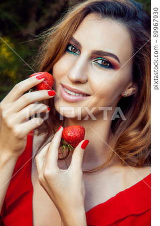 Young beautiful happy funny girl with red dress and makeup holding strawberry in summertime in the park. healthy lifestyle, diet beauty and happiness concept. 89967930