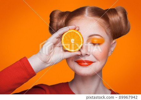 Young beautiful funny fashion model with orange slice on orange background. with orange makeup and hairstyle and freckles. holding orange between eyes with nice smile. Young beautiful funny fashion model with orange slice on orange background. with orange makeup and hairstyle and freckles. holding orange between eyes with nice smile. 89967942