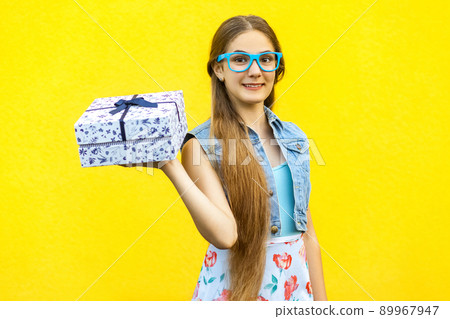 Portrait of a happy hipster smiling girl in dress and blue glasses, holding present box and toothy smile isolated over yellow background. Indoor studio shoot Portrait of a happy hipster smiling girl in dress and blue glasses, holding present box and toothy smile isolated over yellow background. Indoor studio shoot 89967947