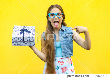 The funny blonde girl in dress and blue glasses, tongue out at the camera, pointing finger on present box, isolated over yellow background. Indoor studio shoot The funny blonde girl in dress and blue glasses, tongue out at the camera, pointing finger on present box, isolated over yellow background. Indoor studio shoot 89967995