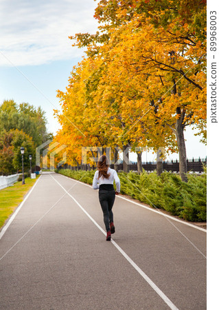 Young adult woman running in autumn forest. Back side view. Girl jogging in road. Outdoor shot 89968003