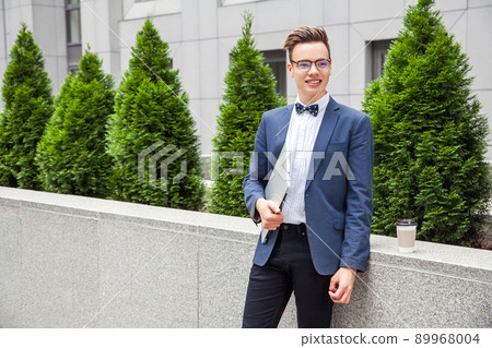 Attractive young businessman with laptop and coffee in hands on office building background. 89968004