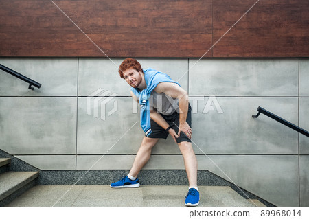 Sport injury. Injured runner, bearded freckled man with athletic body on running track feeling pain of knee. Outdoor summer activity, sport. Healthy lifestyle and workout. 89968014