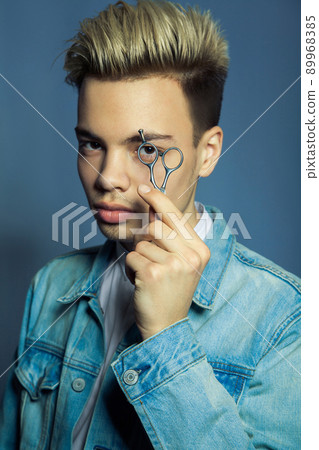 Portrait of fashion blonde Barber hairstylist with scissors in hand near eye on blue background. 89968385