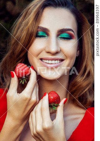 Young beautiful happy funny girl with red dress and makeup holding strawberry in summertime in the park. healthy lifestyle, diet beauty and happiness concept. 89968397